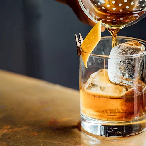 A glass tumbler with whiskey on the rocks, ice cubes catching light, a citrus garnish on the rim, and a bar behind.