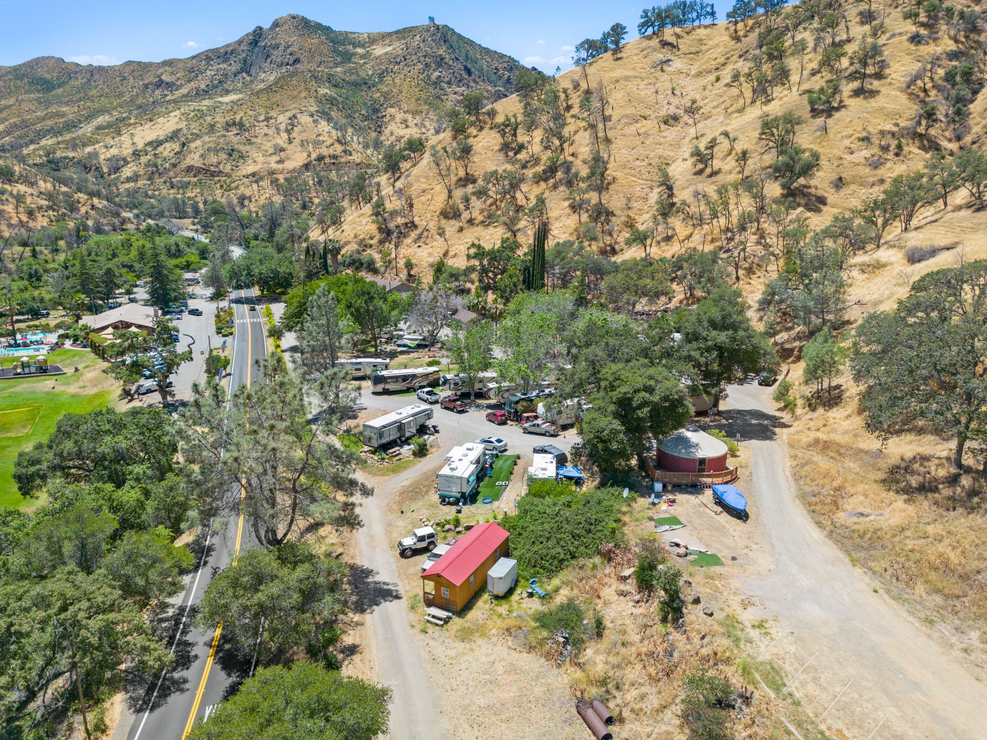 The image shows a rural landscape with roads, buildings, vehicles, and hills in the background under a clear blue sky.