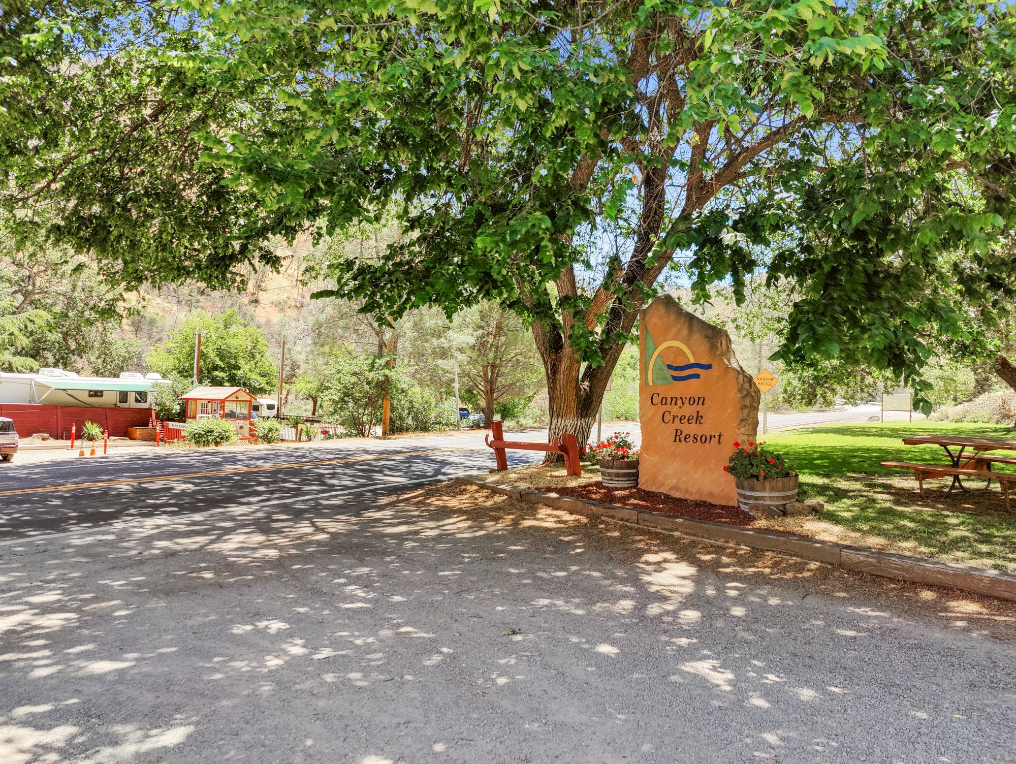 A campsite entrance with a large sign, trees, parked RV, and picnic tables in a sunny outdoor setting.
