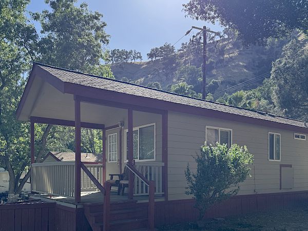 A small, single-story house with a covered front porch, set in a sunny, rural yard with trees and a dirt foreground.