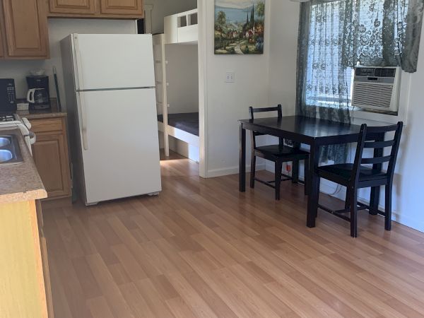 Cozy kitchen and dining area with a white fridge, wooden cabinets, small dining table, and a window with sheer curtains; light hardwood floors.