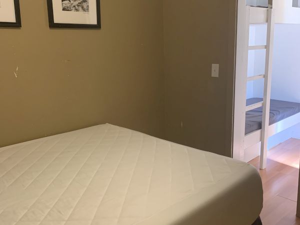 A small bedroom with a queen bed, plain beige walls, two framed pictures, and an open doorway leading to a bright loft area with a bunk or ladder.