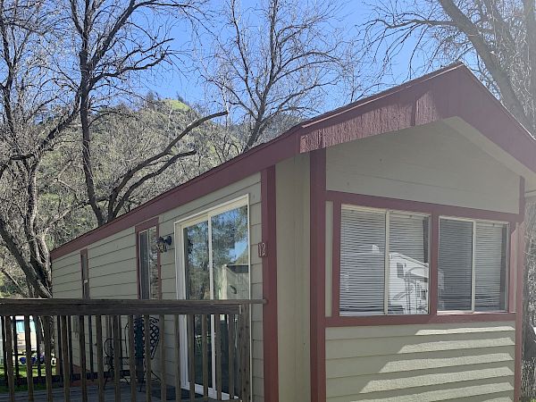 A small cabin with beige siding, red trim, a front porch, and bare trees in the yard under a clear blue sky.