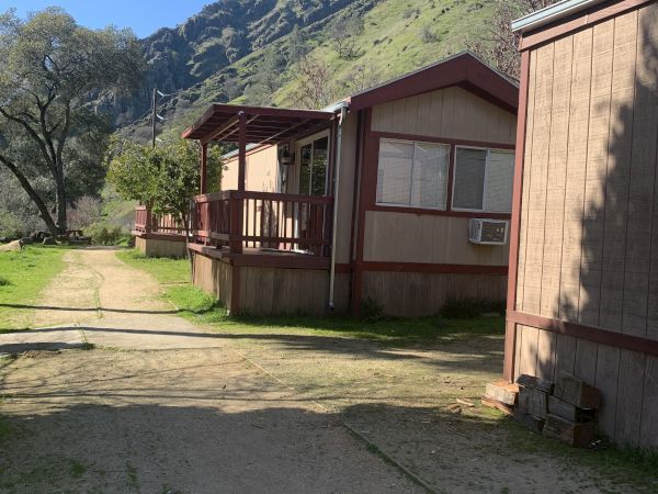 A rural mobile home park with a wooden porch, a dirt road, and green hills/mountains in the background.