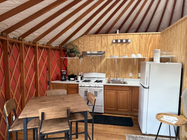 A cozy kitchen setup inside a yurt, featuring a stove, refrigerator, table, and chairs, with wood paneling and a red lattice wall.