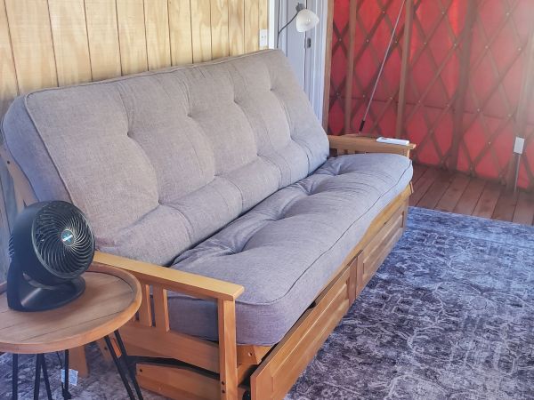 A cozy room with a gray futon, small side table with a fan, and wooden walls. An air conditioner and floor lamp are also visible.
