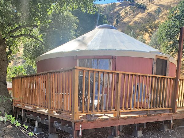The image shows a yurt with a wooden deck surrounded by trees and a hillside in the background.