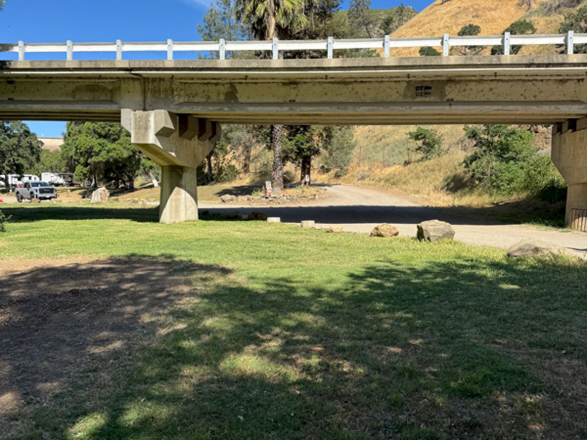The image shows a bridge over a grassy area with trees and a hill in the background under a clear blue sky.