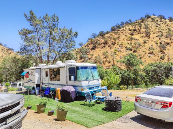An RV is parked in a scenic area with hills. There are chairs, a table, and plants around, creating a cozy outdoor setup.