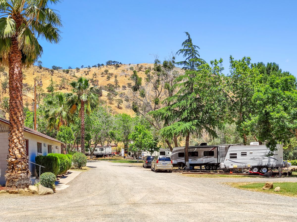 The image shows a rural scene with a house, parked RVs, trees, and a dirt road in a hilly landscape under a clear blue sky.