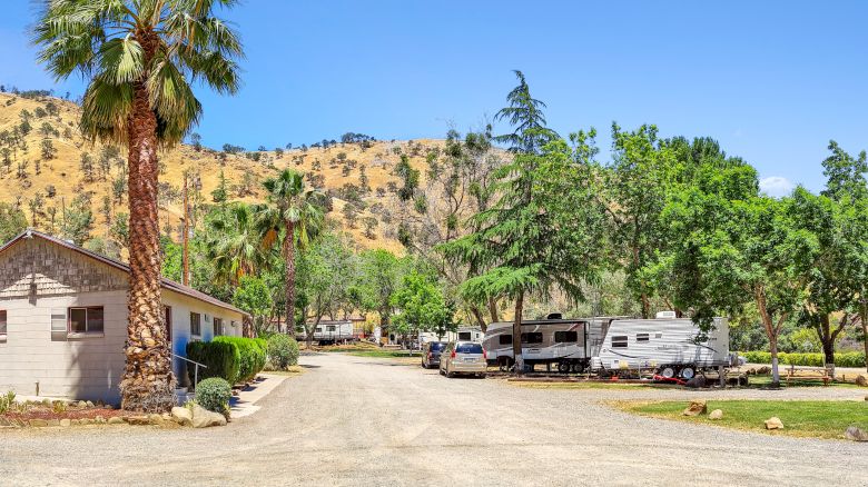 The image shows a rural scene with a house, parked RVs, trees, and a dirt road in a hilly landscape under a clear blue sky.