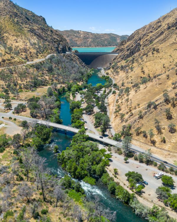 A scenic canyon with a river flowing between steep, rocky hills, a bridge crossing the water, and a winding road lined with trees and greenery.