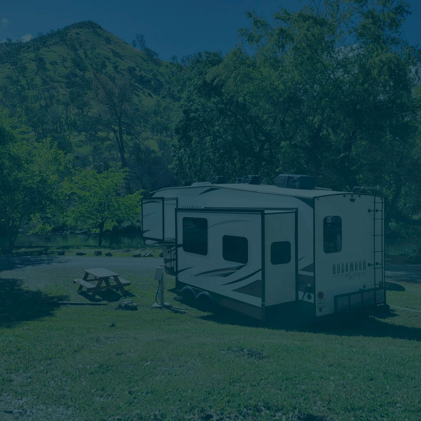 The image shows an RV parked on a grassy area with trees and hills in the background. A picnic table is nearby.