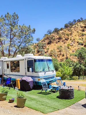 An RV is parked on a green mat with chairs and a fire pit. It’s surrounded by hills and trees under a clear blue sky.
