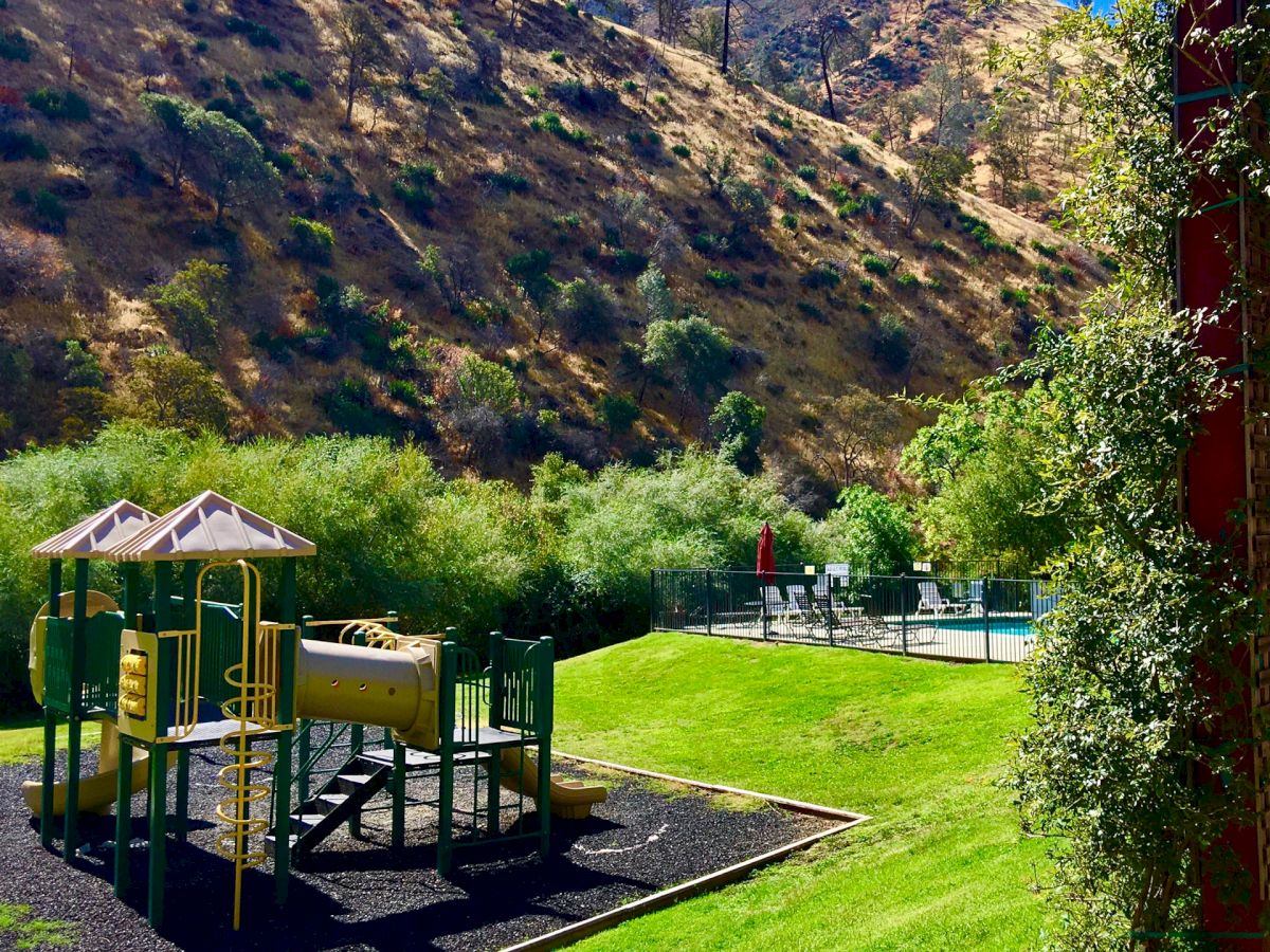 A playground with slides and climbing equipment is set in a grassy area by hills, under a clear blue sky.