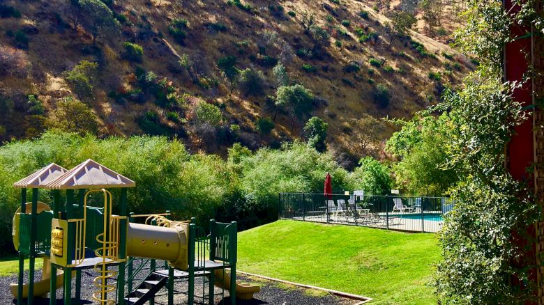 A playground with slides and climbing equipment is set in a grassy area by hills, under a clear blue sky.