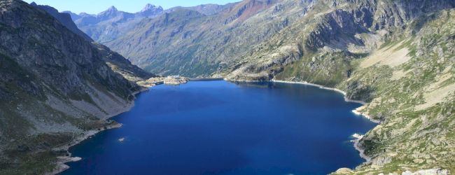 A serene mountain lake surrounded by rugged peaks under a clear blue sky, with patches of green and rocky terrain visible around the water.