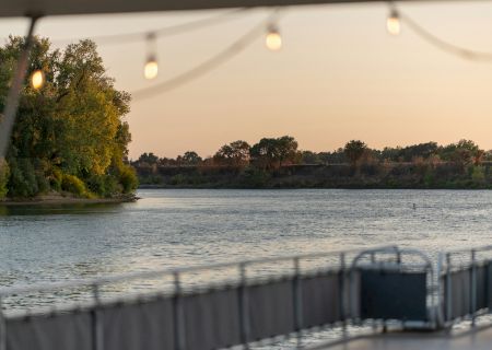 A peaceful riverside scene with trees in the background, gentle lighting, and a railing in the foreground, under a clear evening sky.