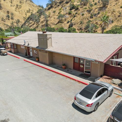 The image shows a modest building with parked cars in a lot, set against a background of dry hills and sparse vegetation.