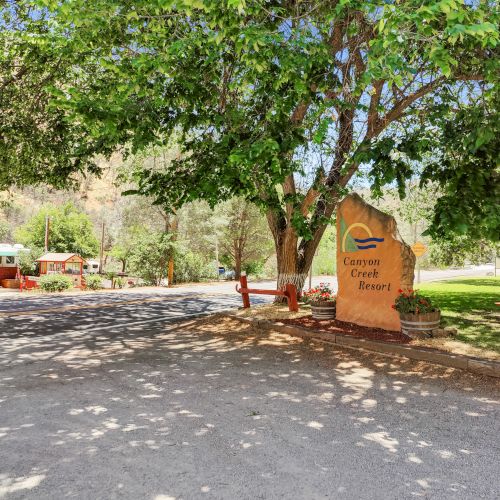 A park entrance with a large sign, trees, benches, and a road in a sunny outdoor setting.