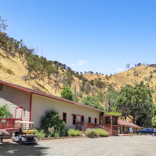 A building flanked by a hilly landscape and trees under a clear blue sky, with golf carts parked nearby in a rural setting.
