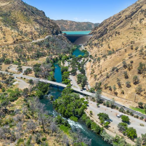A scenic view of a river flanked by hills, with a bridge and a dam in the background under a clear blue sky.