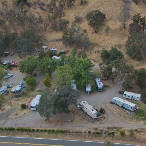 The image shows an aerial view of a campground with several RVs and trees, situated near a road and surrounded by dry landscape.