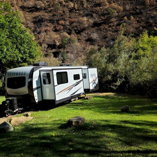A camper is parked on lush green grass surrounded by trees and hills under bright sunlight.