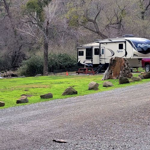 The image shows a parked RV and a red pickup truck on a grassy area with trees in the background, and a gravel path in the foreground.