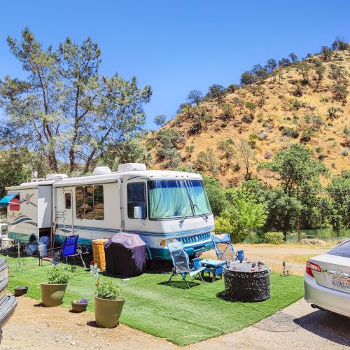An RV parked at a campsite with outdoor chairs, a table, and a fire pit. Surrounding it are hills, trees, and several parked cars.