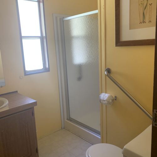 A small bathroom with a shower, toilet, sink, window, and a wall-mounted handrail. The decor is minimal and functional.