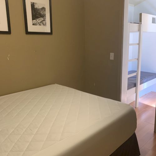 A bedroom with a bed, two framed pictures on the wall, and a view into another room with bunk beds through an open door.