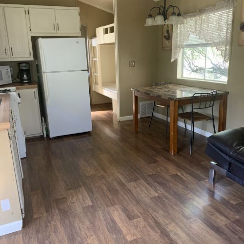 The image shows a small kitchen with a white fridge, a dining table with chairs, a bunk bed in the background, and wooden floors.