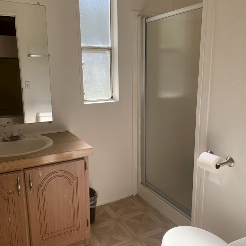 The image shows a bathroom with a sink, mirror, shower, toilet, and a frosted window for privacy, all in a compact space.
