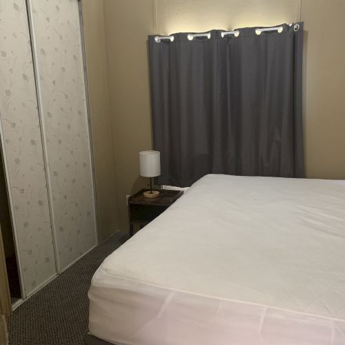 A bedroom with a bed, side table, lamp, patterned closet doors, and gray curtains are visible in the image.