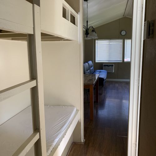 The image shows a narrow room with bunk beds on the left and a living area with a table and chairs at the end of the hallway.
