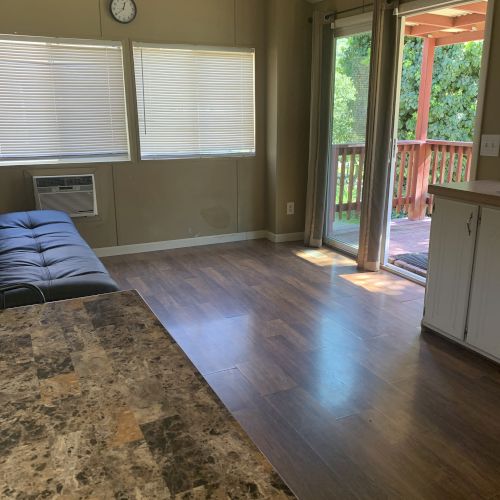 A room with a marble table, couch, windows with blinds, an air conditioner, and an open door to a wooden porch with greenery.