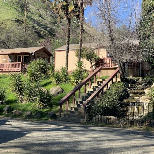 The image shows a rustic scene with wooden cabins, a staircase, palm trees, and a dirt path surrounded by greenery and hills.