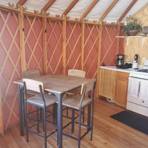 A cozy yurt interior with a wooden dining table, chairs, a kitchenette, and a potted plant, giving a rustic and inviting ambiance.