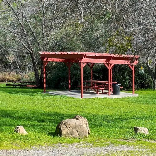A red picnic shelter with a table sits on a grassy area, surrounded by trees and rocks in a scenic outdoor setting.