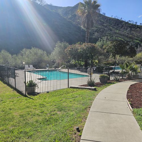 A fenced pool area with lounge chairs, surrounded by greenery and hills under a sunny sky.