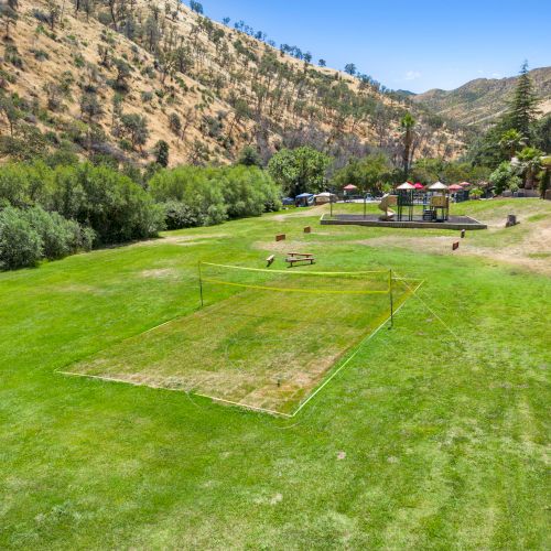 A grassy field with a fenced area, surrounded by hills and trees. Picnic tables and a small structure are in the background.