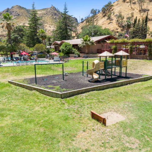 A playground with slides, a pool area, and picnic tables under umbrellas are surrounded by grassy hills and trees in a sunny setting.