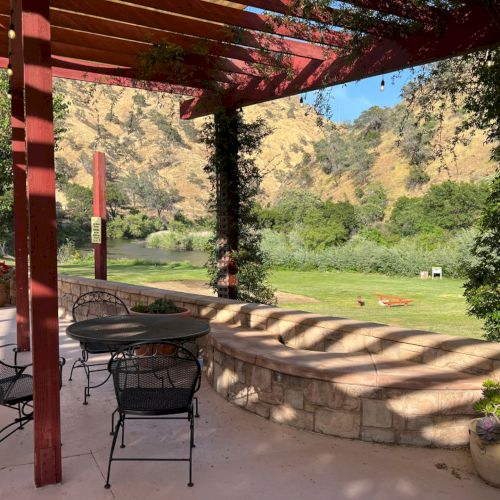 A serene patio with metal chairs and a table under a pergola, overlooking a lush landscape with hills and greenery in the background.