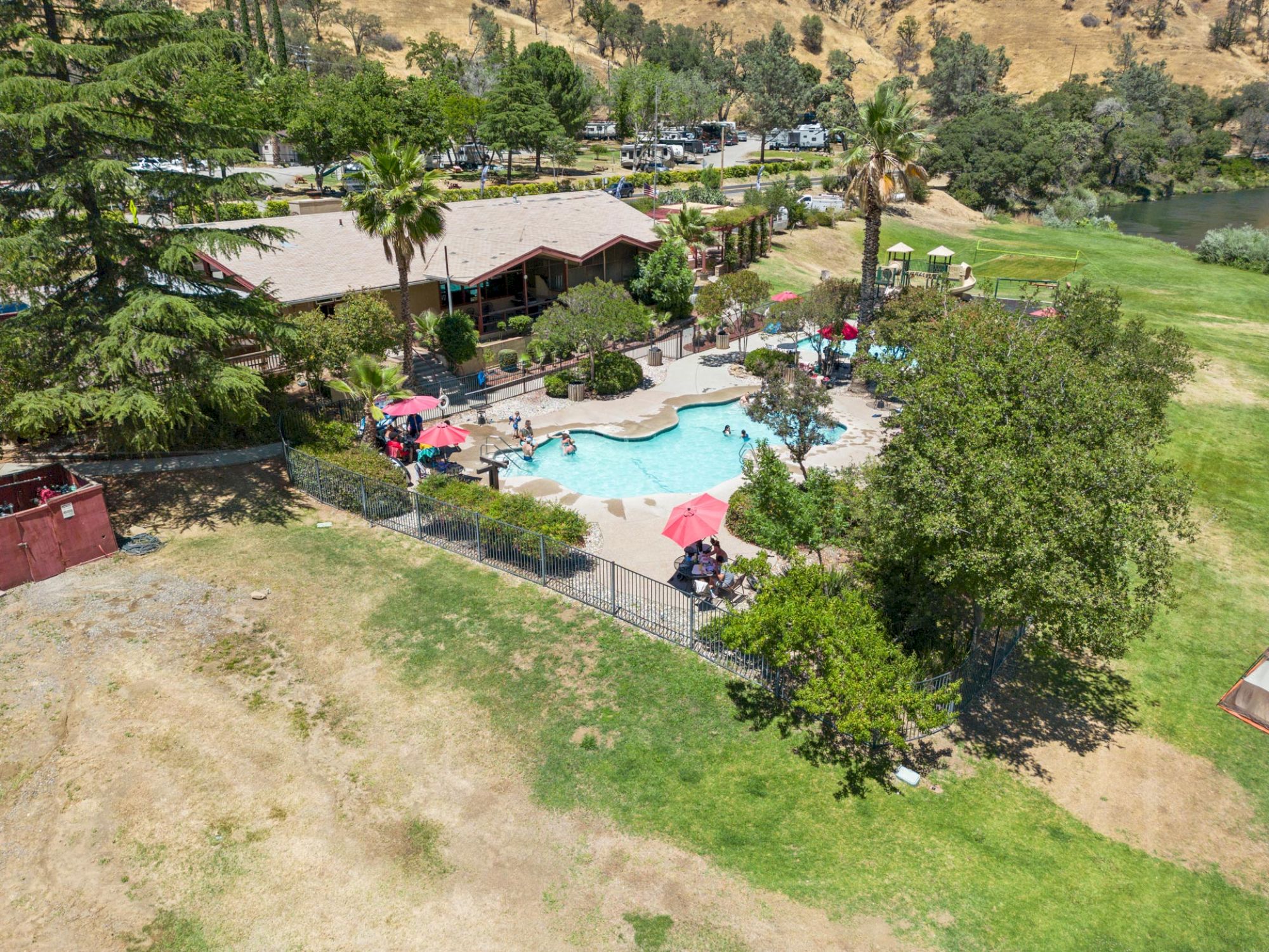 An aerial view of a pool area with loungers and umbrellas, surrounded by trees and a building, set in a hilly landscape.
