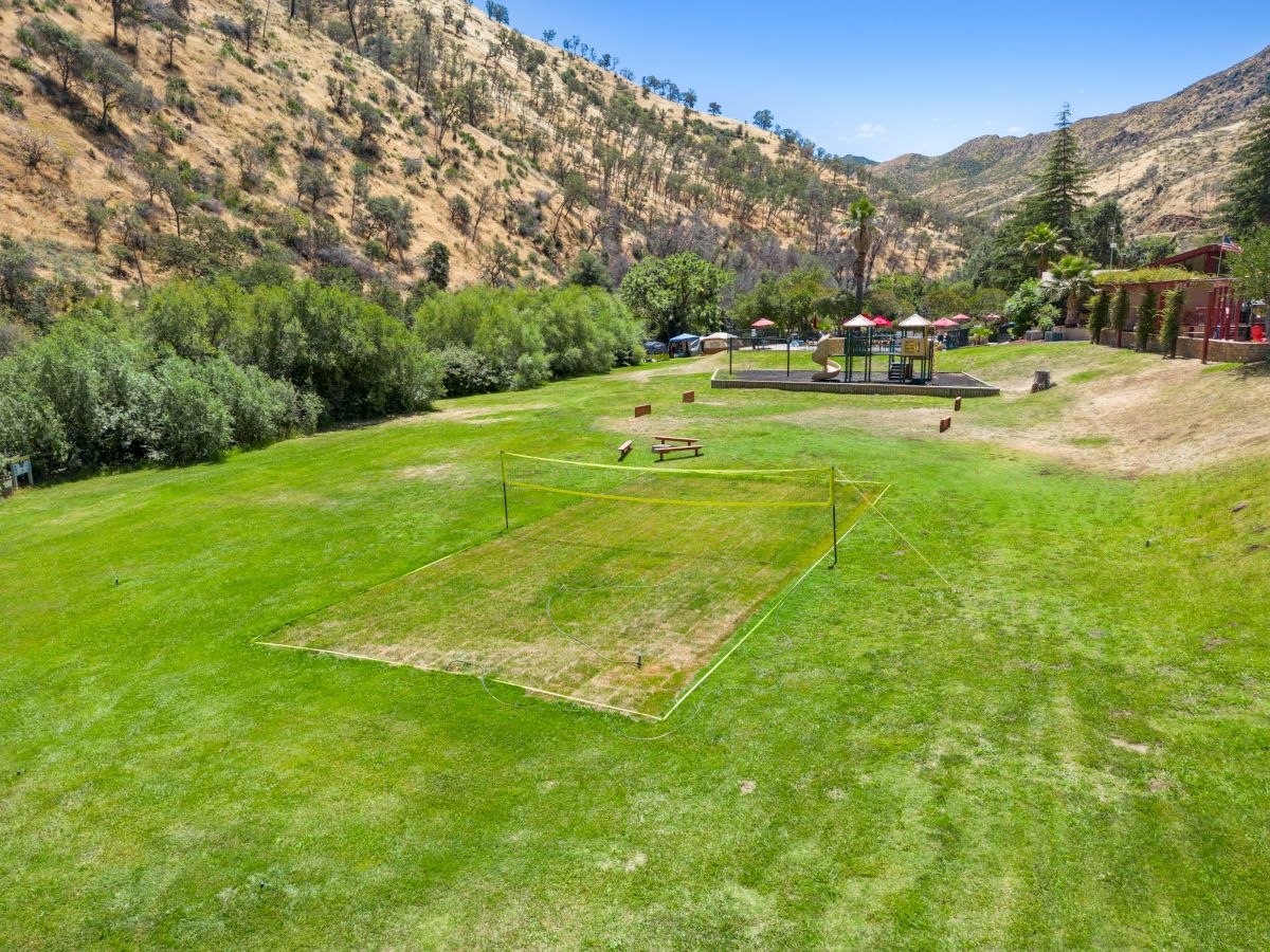 A sunny hillside park with a trimmed grassy area and a rectangular cricket or bocce court in the foreground, trees and benches in the distance, and mountains beyond.