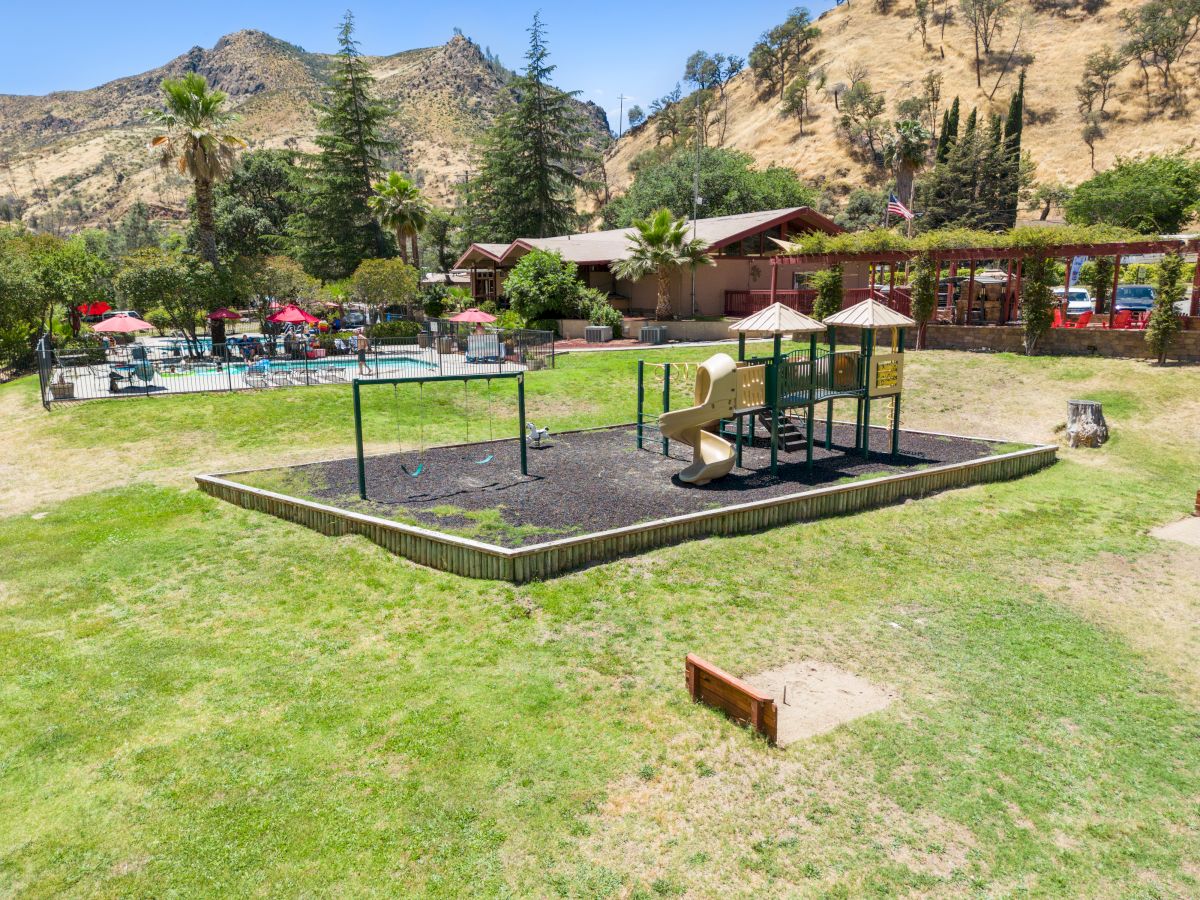 A playground with swings and a small slide sits on a raised platform in a grassy park, mountains and trees in the background, sunny day.