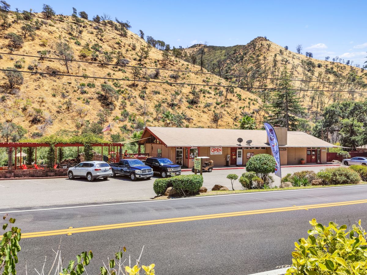 A single-story building with a brown roof sits by a road, nestled against rocky hills, cars parked in front, and a blue sky above.