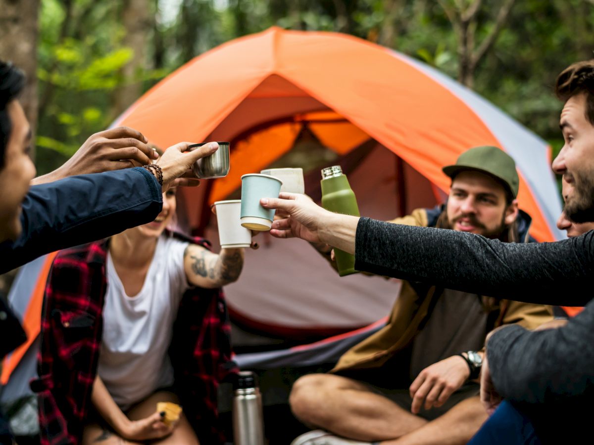 Group of friends camping, toasting cups in front of an orange tent, sharing drinks around a campfire vibe.