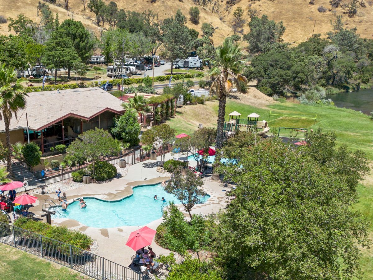 An aerial view of a resort-style pool area with palm trees, a clubhouse, lounge chairs, and red umbrellas, surrounded by green lawns and hills.